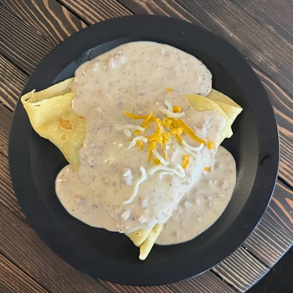 Savory crepes covered in creamy sausage gravy, topped with shredded cheese on a black plate.
