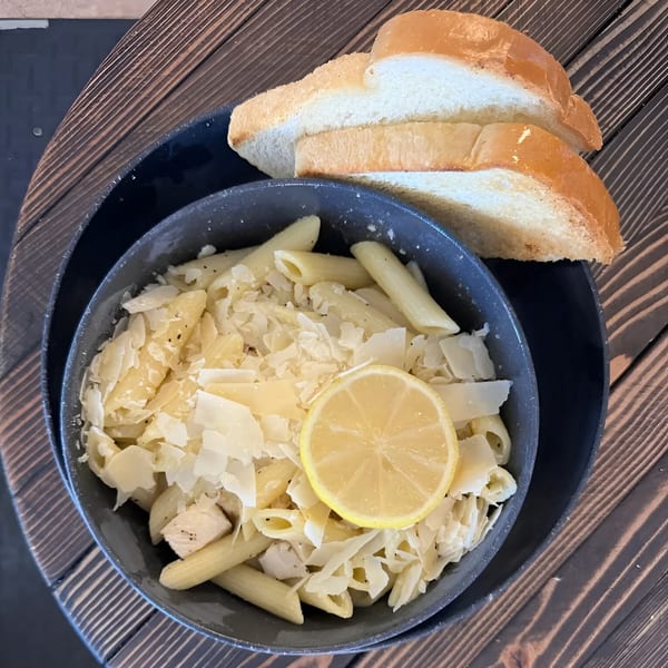 Penne pasta with chicken, shaved Parmesan, and a lemon slice, served with bread on a wooden table.